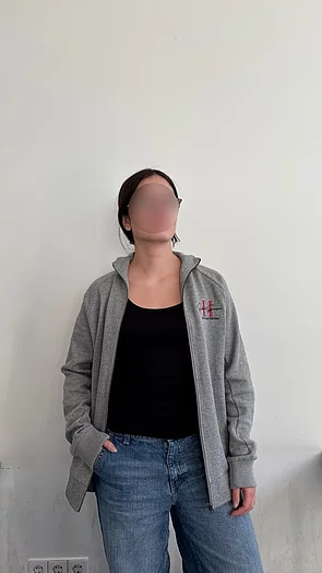 Eine Frau trägt einen grauen Sweatshirt und bluejeans, steht vor einer hellen Wand.