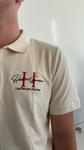 Ein Mann trägt ein beiges Poloshirt mit dem Logo des Haldensleben-Gymnasiums in roten und schwarzen Buchstaben.