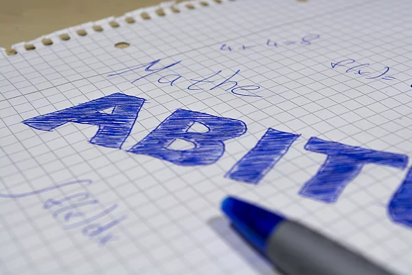 Handgeschriebene Notizen auf kariertem Papier mit dem Wort "ABITUR" in blauer Farbe und einer blauen Kugelschreibermine.