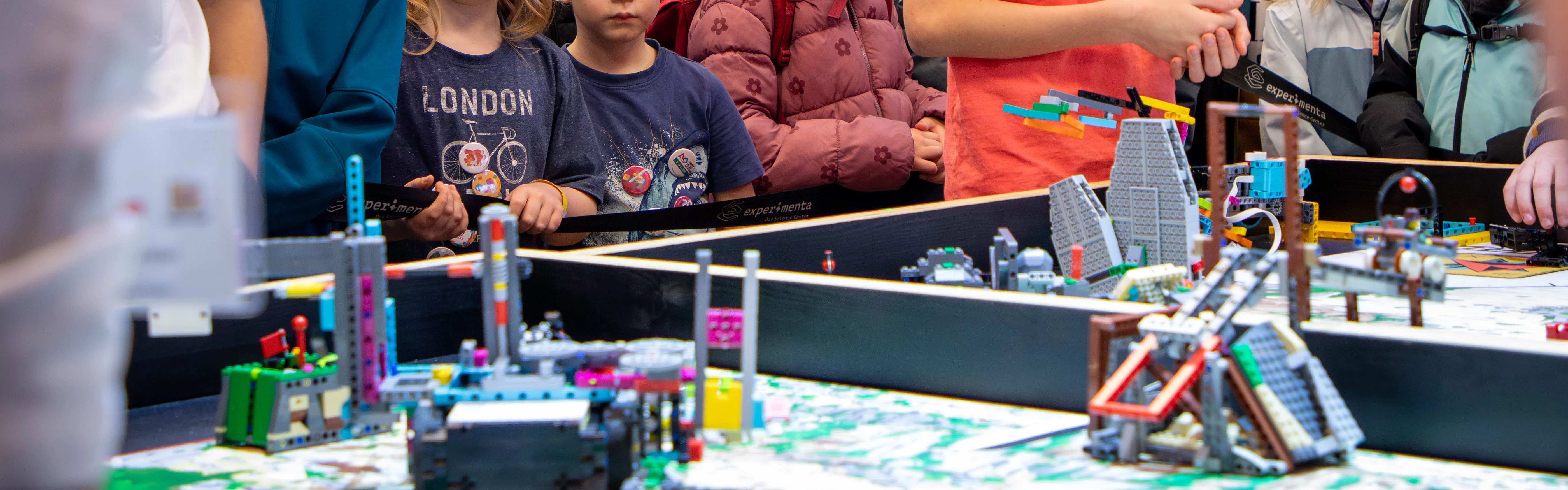 Eine Gruppe von Kindern und Erwachsenen beobachtet einen Roboter-Wettbewerb mit Lego-Modellen.