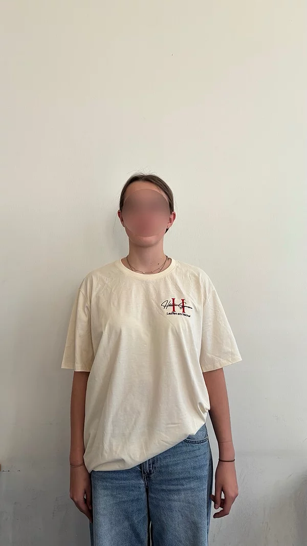 Eine Person steht vor einer neutralen Wand, trägt ein beiges T-Shirt und Jeans.