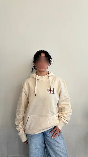 Eine Person trägt einen hellbeigen Kapuzenpullover und Jeans, vor einem neutralen Hintergrund.