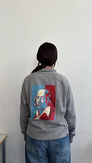 Eine Person von hinten trägt einen grauen Pullover mit einem farbenfrohen Porträt auf dem Rücken.