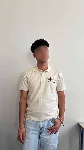 Junger Mann steht vor einer weißen Wand, trägt ein helles Polohemd und Jeans. Hände leicht verschränkt.