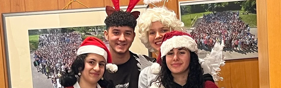 Vier Personen in festlicher Kleidung posieren am Tisch; zwei als Weihnachtsmann, eine als Engel und eine als Rentiere.