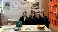 Drei junge Frauen sitzen an einem Tisch mit Informationen zur Eine-Welt-AG und einem Korb mit "Lose".